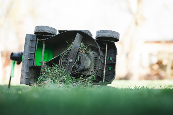 A gas-powered lawnmower is shown on its side, revealing the dirty blades and a buildup of grass clippings underneath. The background is a blurry outdoor scene with a house.